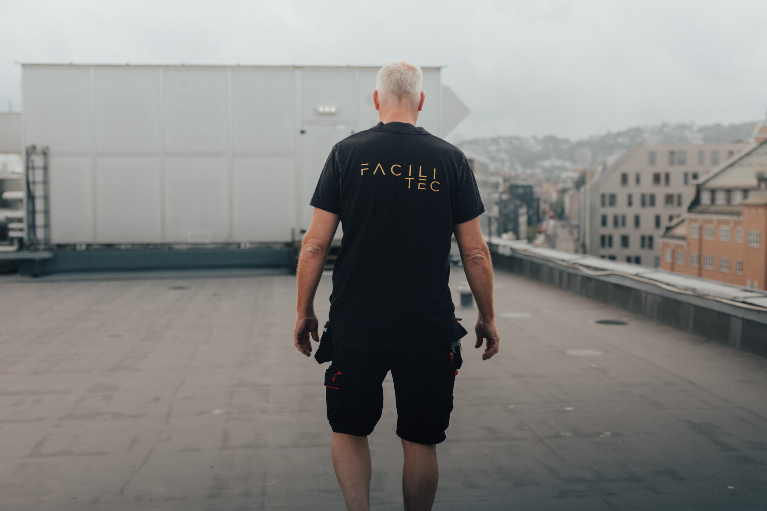 En man står med ryggen mot kameran på ett tak mitt i staden med lägenhetshus i bakgrunden en grå dag. Foto
